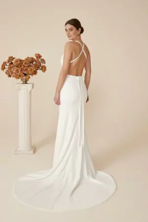 Robe de mariée ivoire à décolleté plongeant, dos nu croisé, coupe ajustée et fente frontale, avec une traîne fluide ornée de boutons.